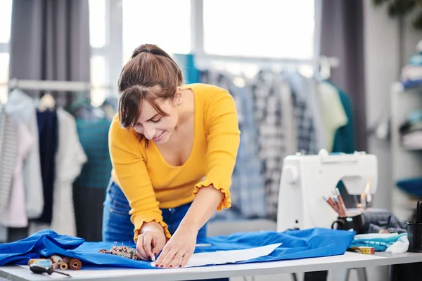 Creative caucasian fashion designer standing in her studio and drawing scheme on blue linen for a beautiful evening dress. — Stock Photo, Image