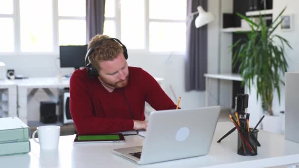 Młody Przystojny Innowacyjny Biały Freelancer Siedzi Jego Biurze Słuchawki Oglądając — Wideo stockowe