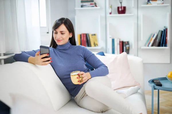 年轻迷人的黑发美女坐在客厅的沙发上，端着杯子喝咖啡，挂在社交媒体上. — 图库照片