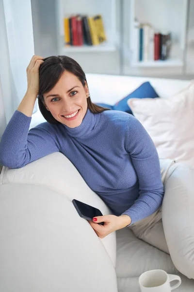 Junge attraktive lächelnde attraktive Brünette sitzt auf der Couch in ihrem Wohnzimmer und entspannt. Sie hält ihr Smartphone in der Hand und blickt in die Kamera. — Stockfoto