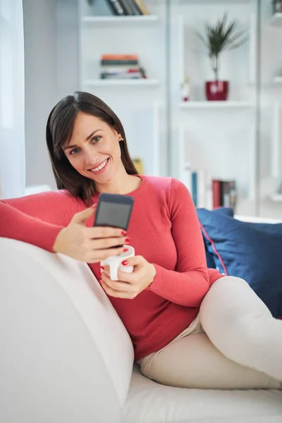 Junge attraktive lächelnde Brünette sitzt auf der Couch in ihrem Wohnzimmer, hält Tasse mit Kaffee und hängt in den sozialen Medien. — Stockfoto