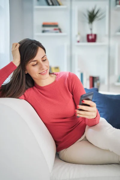 Junge hinreißende Brünette sitzt auf Sofa in ihrem Wohnzimmer und nutzt Smartphone zum Internetsurfen. — Stockfoto
