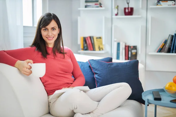 Morena Atraente Sentada Sofá Com Pernas Cruzadas Lendo Livro Bebendo — Fotografia de Stock