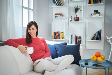 Çekici esmer kanepede bacak bacak üstüne atmış kitap okuyor, kahve içiyor ve dinleniyor..