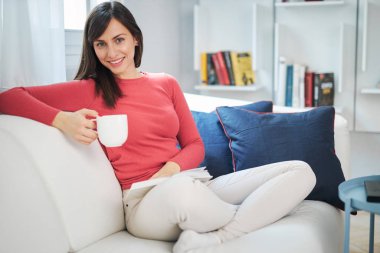 Çekici esmer kanepede bacak bacak üstüne atmış kitap okuyor, kahve içiyor ve dinleniyor..