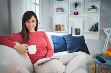 Çekici esmer kanepede bacak bacak üstüne atmış kitap okuyor, kahve içiyor ve dinleniyor..