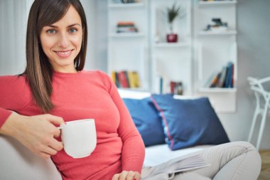 Çekici esmer kanepede bacak bacak üstüne atmış kitap okuyor, kahve içiyor ve dinleniyor..