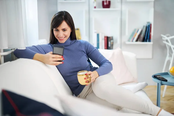 年轻迷人的黑发美女坐在客厅的沙发上 端着杯子喝咖啡 挂在社交媒体上 — 图库照片