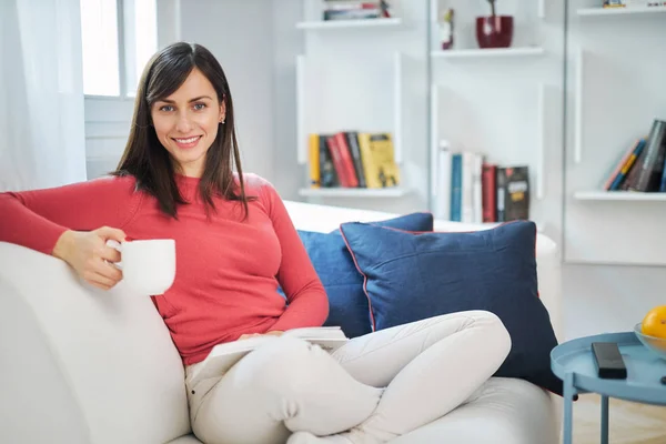 Morena Atraente Sentada Sofá Com Pernas Cruzadas Lendo Livro Bebendo — Fotografia de Stock