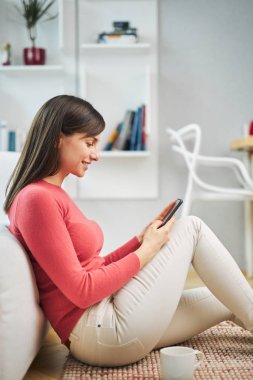 Genç, çekici, gülümseyen bir kadın evde yerde oturuyor, akıllı telefon kullanıyor ve dinleniyor..