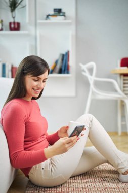 Genç, çekici, gülümseyen bir kadın evde yerde oturuyor, akıllı telefon kullanıyor ve dinleniyor..