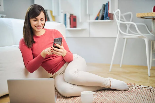 年轻的微笑的女人坐在家里的地板上 用智能手机 — 图库照片