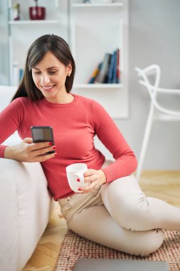 Genç, çekici, gülümseyen bir kadın evde yerde oturuyor, akıllı telefon kullanıyor ve dinleniyor..