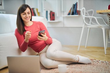 Genç, gülümseyen bir kadın yerde oturuyor ve akıllı telefon kullanıyor..