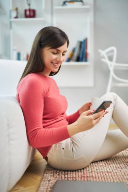 Genç, gülümseyen bir kadın yerde oturuyor ve akıllı telefon kullanıyor..