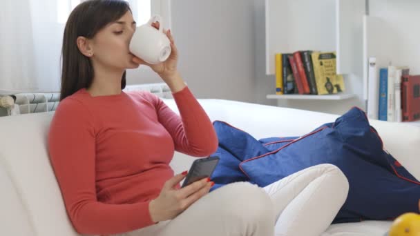 漂亮的黑发女人坐在她家的沙发上 用智能手机打字 喝咖啡 — 图库视频影像