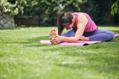 Spor kıyafetli genç esmer arka bahçesindeki paspasın üzerinde oturuyor ve bacağını esnetiyor..