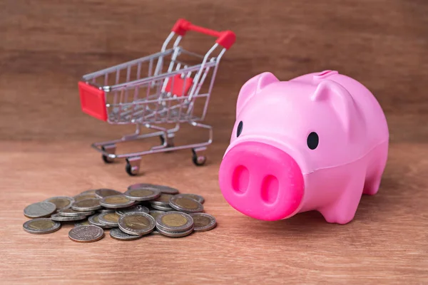 Alcancía de plástico con monedas y carrito de compras en la mesa de madera — Foto de Stock