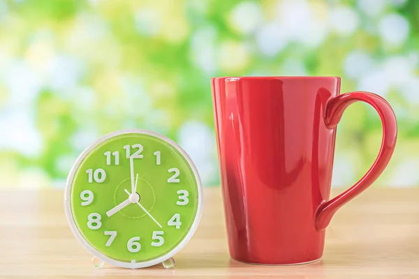 Red coffee cup and clock on wooden table with green leaves bokeh — Stock Photo, Image