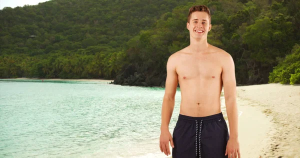 Portrait of attractive guy standing shirtless in swim shorts on Caribbean beach.