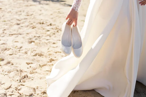 Wedding warm white shoes in brides hands on the beach. — Stock Photo, Image