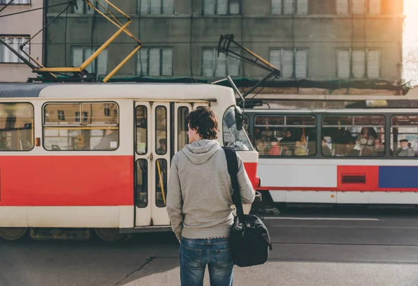 Yol tramvay ile önünde dostum. Çanta ile rahat kıyafetler erkek turist önünde duruyor yol tramvay ile güneşli yaz günü geçiyor. Genç adam bir tren ile bir yolculuk için hazır.