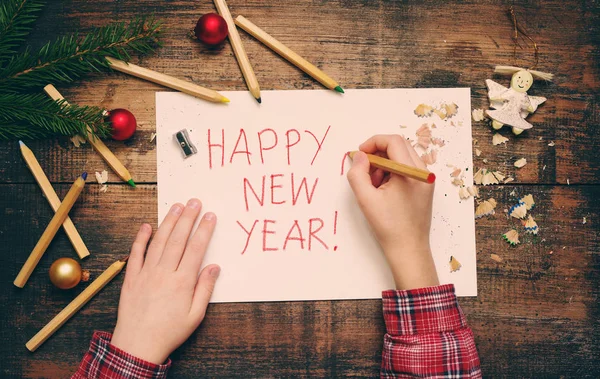 Il bambino scrive "Happy New Year" su un foglio di carta bianco. Mani del bambino, il foglio di carta, matite e decorazioni di Natale — Foto Stock