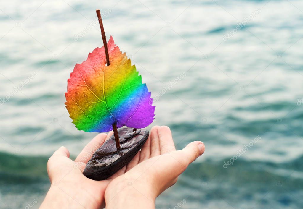 Small decorative sailboat with a rainbow a sail in LGBT colors in the hands against the background of the sea. 