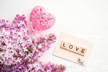 Text LOVE made from wooden letters, pink heart and lilac flowers on a white wooden table surface, close up, selective focus. Romantic background.