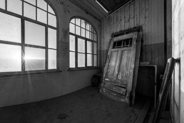 Gün doğumunda terk edilmiş bir evin içinde dramatik bir siyah beyaz fotoğraf, kırık camlardan güneş ışığı ve çöl kumuna gömülü bir kapı, Namibya 'nın hayalet kasabası Kolmanskop' ta çekildi..