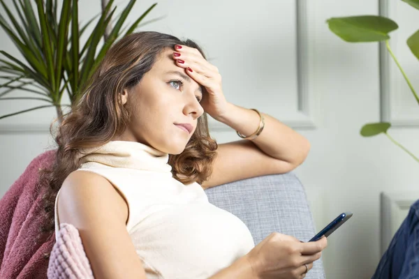 Traurige junge Frau sitzt auf Sofa und liest Telefonnachricht — Stockfoto