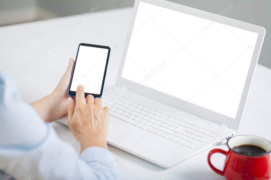 Woman holding a white screen smartphone and blank white screen laptop computer on a desk. Template for your designs easy editable