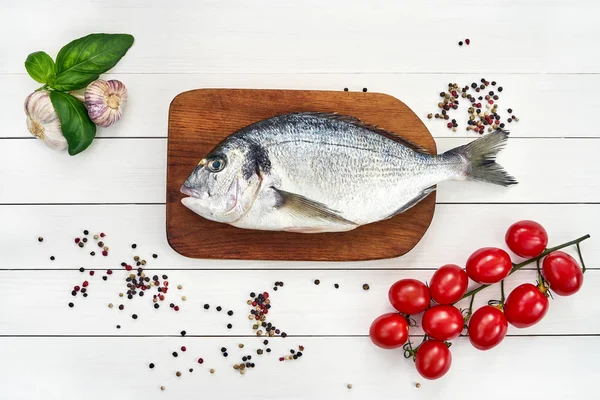 Dorado poisson frais sur planche à découper en bois avec ail, tomates et grains de poivre . — Photo