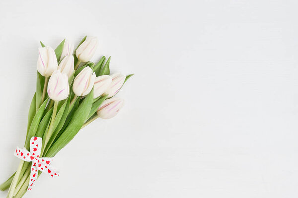 White tulips bouquet decorated with hearts ribbon on white wooden background. Copy space, top view. Valentines Day concept