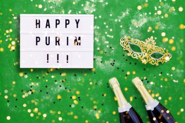 Işık kutusunda mutlu Purim yazıları, iki şampanya şişesi, karnaval maskesi ve yeşil arka planda konfeti. Purim Karnavalı 'nın düz yatışı konsepti. Metin için alanı kopyala.