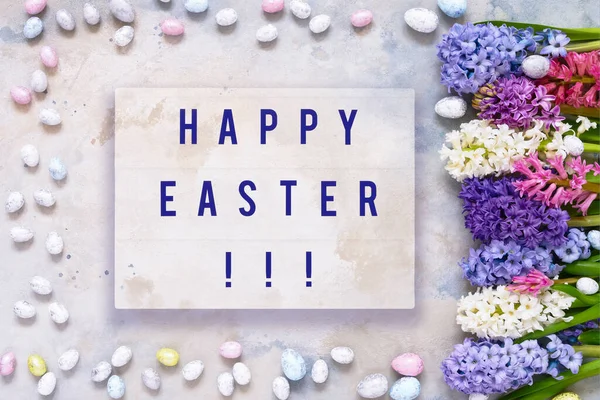 Easter background. HAPPY EASTER written in light box, decorative Easter eggs and hyacinth flowers flatlay. Copy space, top view. Easter celebration concept.