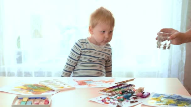 Niño de pelo rubio se sienta a la mesa y comienza a dibujar . — Vídeo de stock