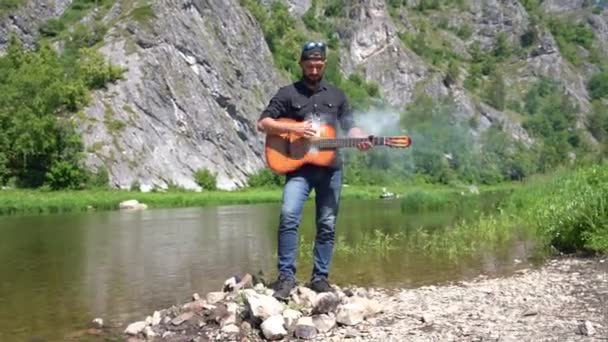 De muzikant speelt gitaar wat de rook is, tegen de achtergrond van de rivier — Stockvideo