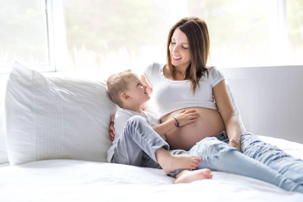 Schöne schwangere junge Mutter, die mit ihrem älteren Kind im Bett sitzt — Stockfoto