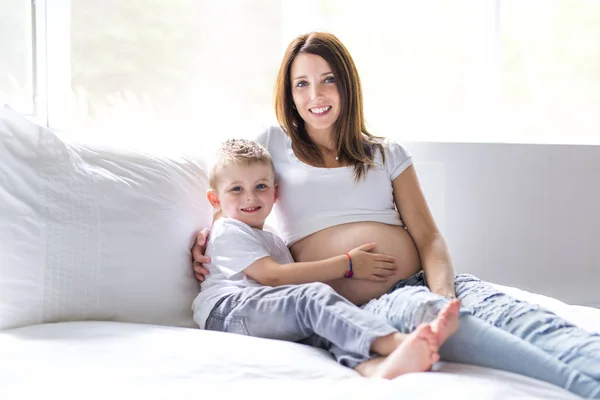 Schöne schwangere junge Mutter, die mit ihrem älteren Kind im Bett sitzt — Stockfoto