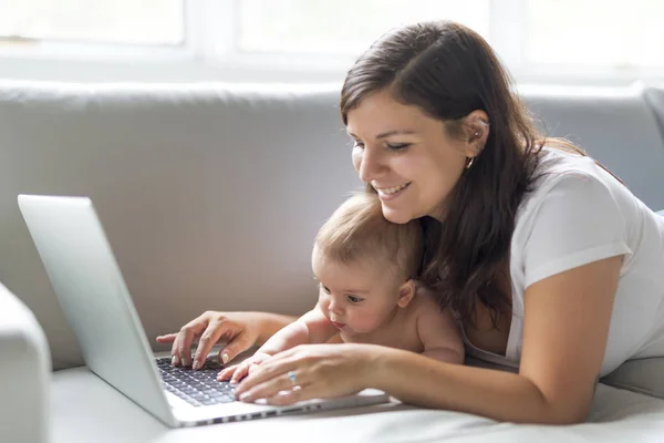 母親と赤ちゃんの子を探している再生し、自宅のソファの上のコンピューターを読む — ストック写真