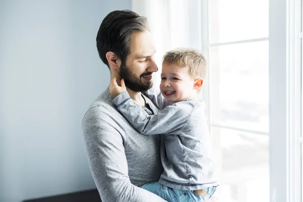 Feliz padre joven sosteniendo chico lindo de pie cerca de la ventana grande . — Foto de Stock