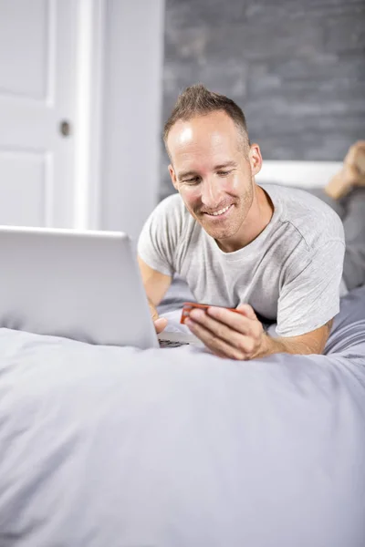 Poważne dorywczo młody człowiek korzysta z laptopa w łóżku w domu — Zdjęcie stockowe