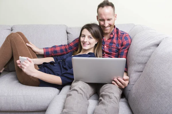 Milující se mladý pár sedí na gauči doma s mobil a laotop — Stock fotografie
