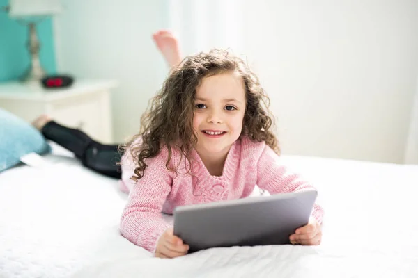 技術は容易になります。デジタル タブレットを保持し、ながらベッドで横になっている笑顔かわいい女の子. — ストック写真
