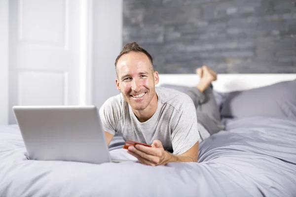 Poważne dorywczo młody człowiek korzysta z laptopa w łóżku w domu — Zdjęcie stockowe