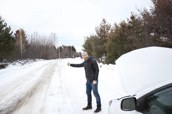 Junger schöner Mann in schwarzer Jacke im Winter, stand auf der Autobahn und trampte. — Stockfoto