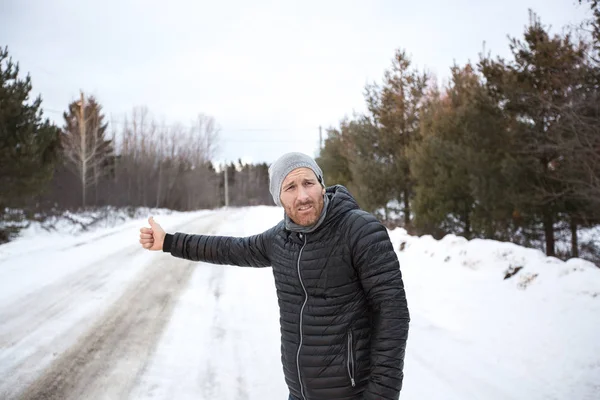 Junger schöner Mann in schwarzer Jacke im Winter, stand auf der Autobahn und trampte. — Stockfoto