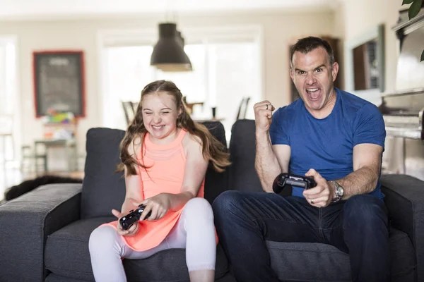 Komea isä ja hänen söpö pieni tyttärensä pelaavat pelikonsolia ja hymyilevät istuessaan sohvalla kotona. . — kuvapankkivalokuva
