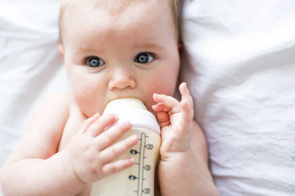 Jolie petite fille boit de l'eau du biberon couché sur le lit. Couche portée enfant dans la chambre d'enfant . — Photo